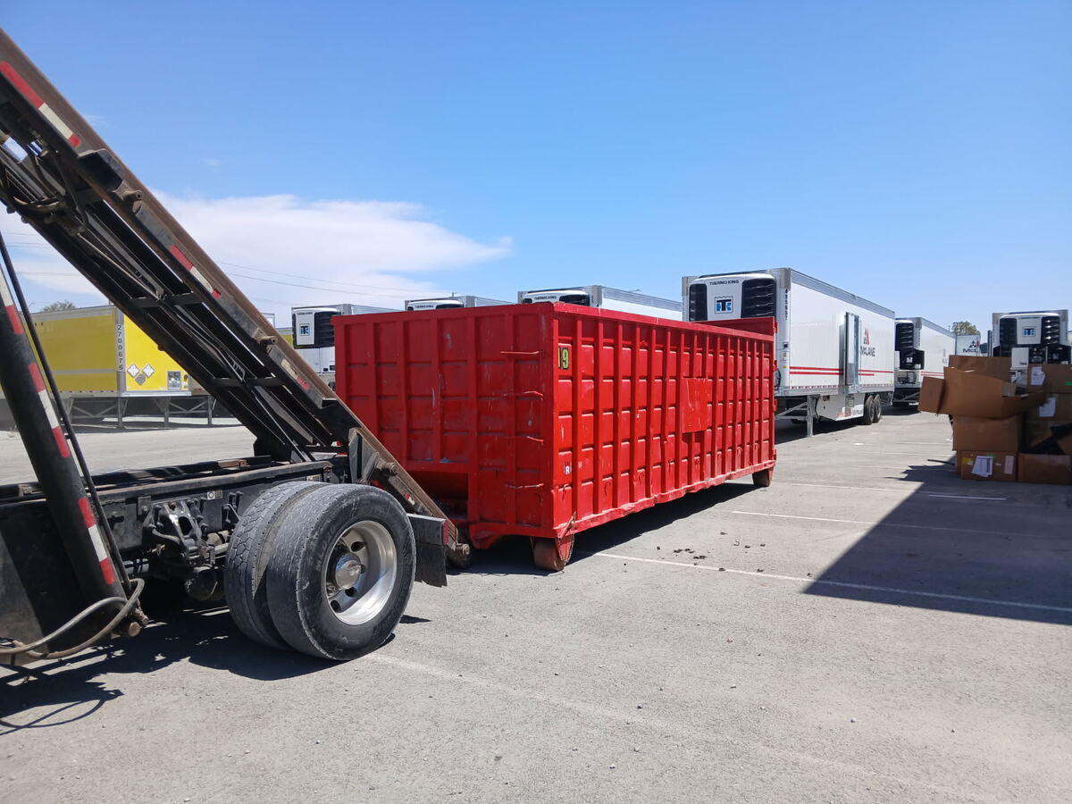 Red 40-yard Heverly & Sons roll-off dumpster being swapped at an Otay Mesa distribution yard next to McLane refrigerated trailers